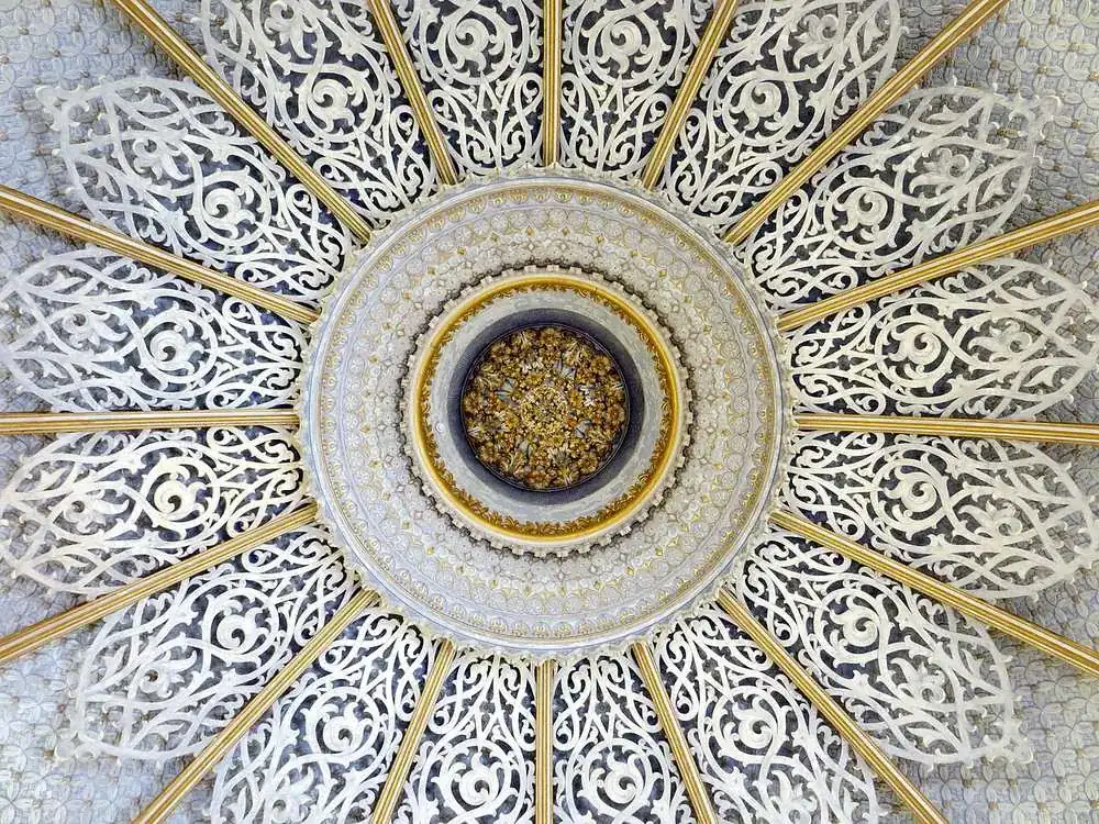 A ceiling seen from directly below — sixteen pierced white ribs radiating from a gilded central boss